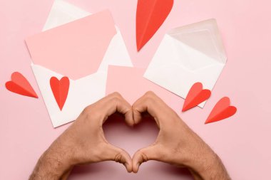 Erkek eli, zarf ile kalp şekli, boş kart ve pembe arka planda kırmızı kağıt kalp yapıyor. Sevgililer Günü kutlaması