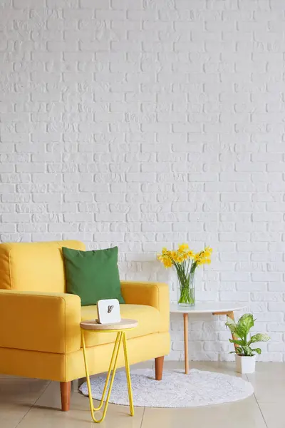 Cozy yellow armchair and vase with blooming narcissus flowers on coffee table near white brick wall