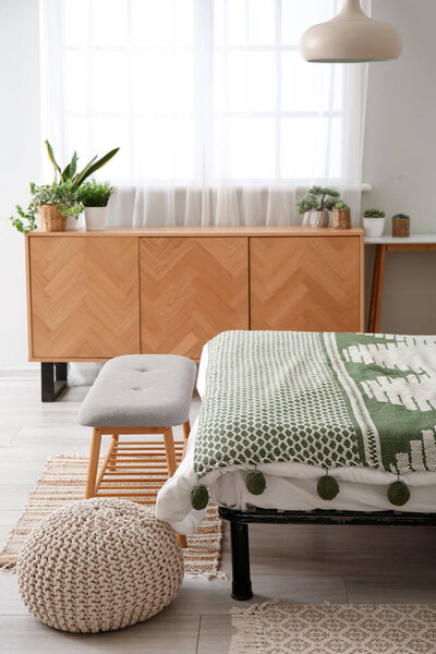 Interior of light bedroom with cozy bed, bedside bench and wooden cabinet near window