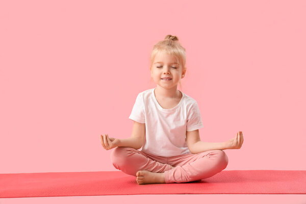 Милая маленькая девочка сидит в позе лотоса на коврике для йоги на розовом фоне