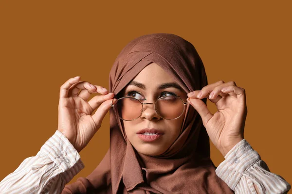 Young African-American Muslim woman in stylish sunglasses with beautiful makeup on brown background