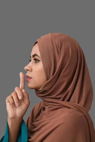 Young African-American Muslim woman with beautiful makeup showing silence gesture on grey background