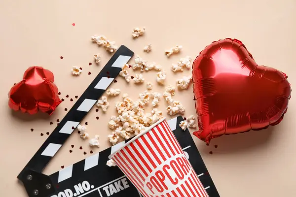 Bucket of popcorn with movie clapper and heart shaped balloons on beige background. Valentine's Day celebration