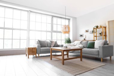 Büyük pencerenin yanında gri kanepeleri ve kahve masası olan aydınlık oturma odasının içinde.