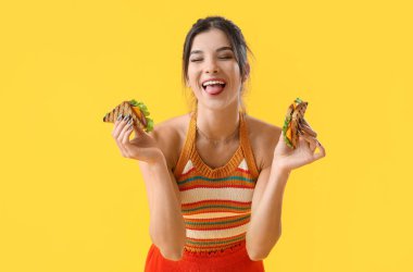 Sarı arka planda dilini gösteren lezzetli sandviçleri olan genç bir kadın.