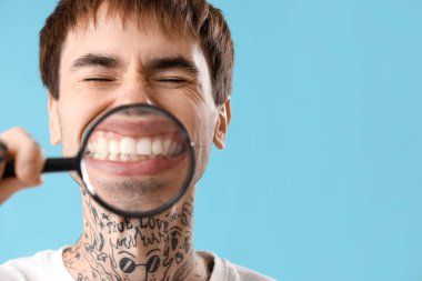Genç dövmeli adam mavi arka planda dişlerini büyütüyor, yakın plan.