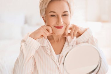 Olgun kadın yatak odasındaki aynanın yanında yüz geliştirme egzersizi yapıyor.