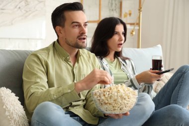 Evde patlamış mısır ve kola içen genç bir çift televizyon izliyor.
