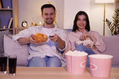 Patates cipsi ve patlamış mısırlı genç çift akşam evde televizyon izliyor.