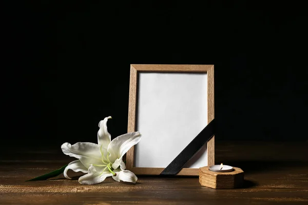 Blank funeral frame, burning candle and lily flower on wooden table against dark background