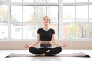Spor salonunun penceresinin yanındaki yoga minderinde meditasyon yapan genç bir kadın.