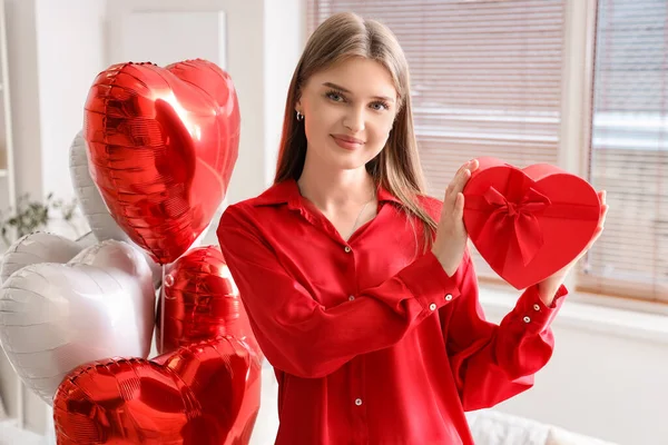 Beautiful woman with gift box and balloons at home on Valentine\'s Day