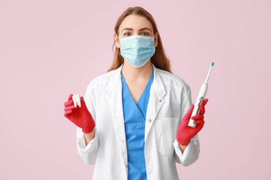 Maskeli, elektrikli diş fırçası ve pembe arka planda diş modeli olan kadın bir dişçi. Dünya Diş Hekimi Günü