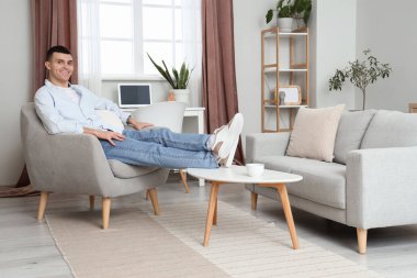 Genç adam koltukta dinleniyor. Ayakları evde masanın üstünde.