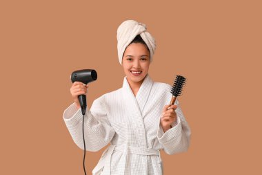 Bornozlu, saç kurutma makinesi ve kahverengi arka planda fırçası olan güzel bir Asyalı kadın.