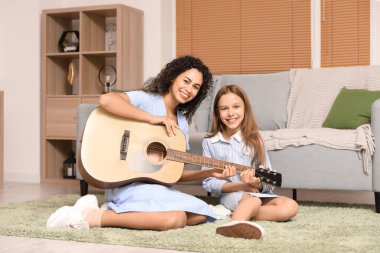 Genç bir kadın, evde yerde otururken akustik gitar çalmayı öğretiyor.