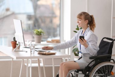 Tekerlekli sandalyedeki genç iş kadını ofiste bilgisayar ve cep telefonuyla çalışıyor.