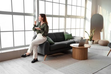 Oturma odasındaki pencerenin yanındaki kanepede oturan bir fincan kahveyle genç bir kadın.