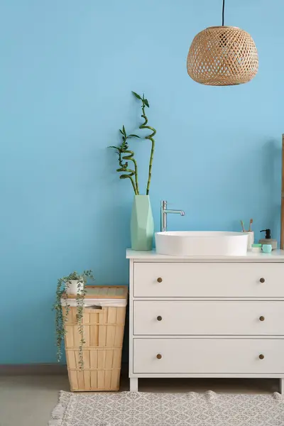 Sink, bath accessories and vase with bamboo stems on drawers near blue wall