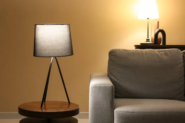Glowing lamp on table near grey sofa in dark room