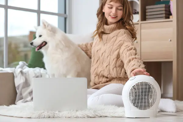 Young woman with Samoyed dog and laptop warming near electric fan heater at home, closeup