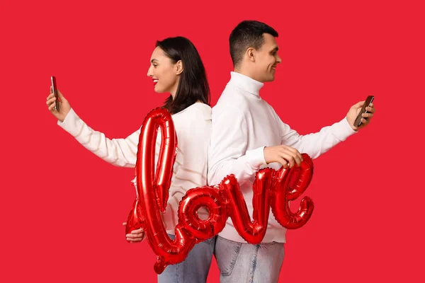 Young couple with mobile phones and air balloon in shape of word LOVE on red background. Online dating