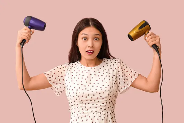 Shocked young Asian woman with hair dryers on pink background