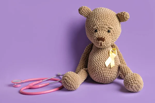 Toy bear with golden ribbon and stethoscope on lilac background. Childhood cancer awareness concept