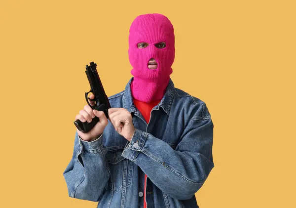 Handsome young man in balaclava with gun on yellow background