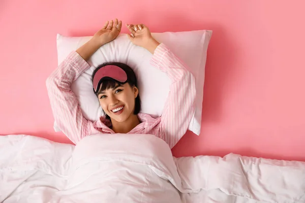 Happy young woman in pajamas with soft pillow and blanket lying on pink background