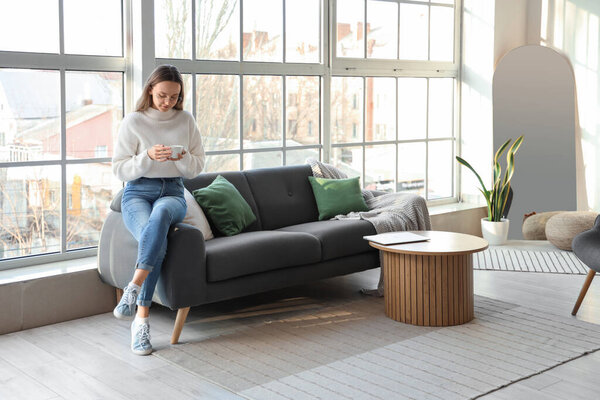 Young woman with cup of tea sitting on sofa near window in living room