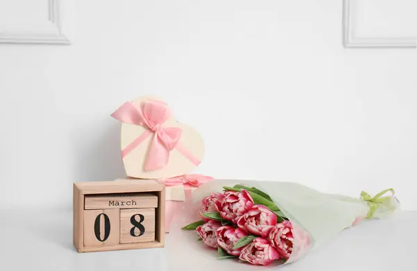 Calendar with date of International Women's Day, tulips and gifts on table near light wall