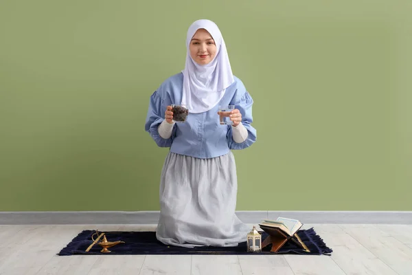 Young Muslim woman with dates and glass of water on mat near green wall. Ramadan celebration