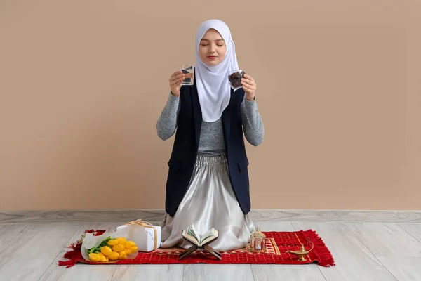 Young Muslim woman with dates and glass of water on mat near beige wall. Ramadan celebration