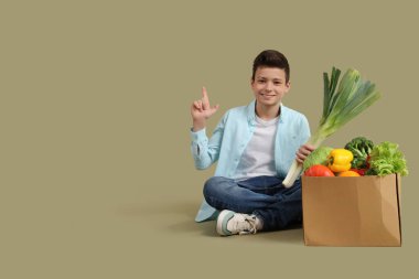 Şirin küçük çocuk yeşil arka planda farklı sebzelerle dolu bir kutuya ve bir şeye işaret ediyor.