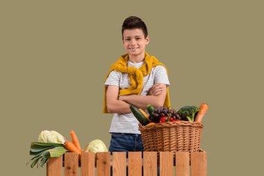 Yeşil arka planda farklı sebzelerle şirin küçük bir çocuk ve hasır sepeti.