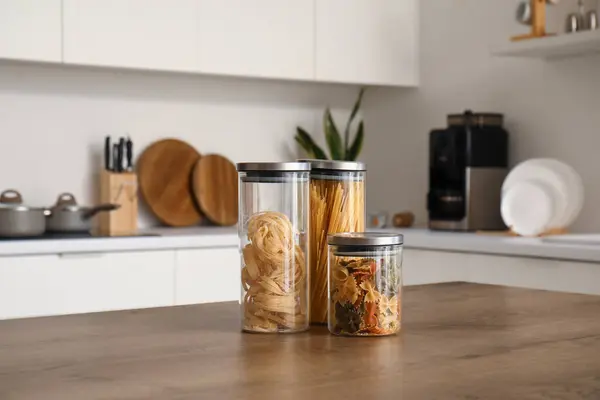 Jars with different types of pasta on wooden table in modern kitchen