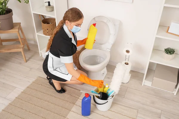 Young chambermaid with medical mask cleaning toilet bowl in bathroom