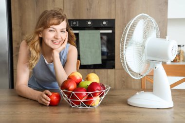 Mutfakta elektrikli yelpaze ve bir kase taze elmayla güzel, mutlu genç bir kadın.