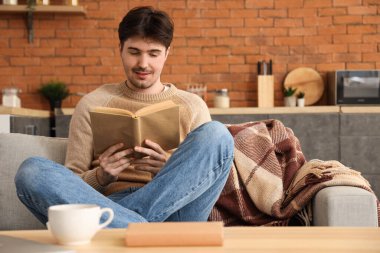 Genç adam evdeki kanepede kitap okuyor.