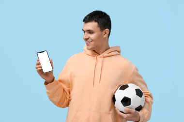 Mavi arka planda futbol topu ve cep telefonu olan genç bir adam.