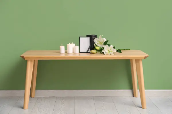 Blank funeral frame, burning candles and beautiful lily flowers on wooden table near color wall