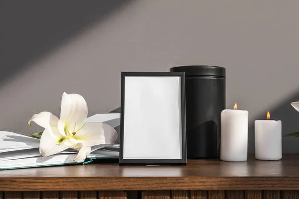 Blank funeral frame, burning candles, mortuary urn and lily flower on wooden cabinet against grey wall