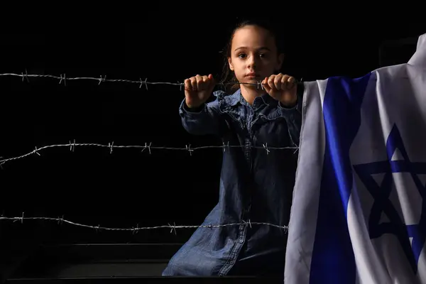 Little Jewish girl with flag of Israel behind barbed wire on black background. International Holocaust Remembrance Day