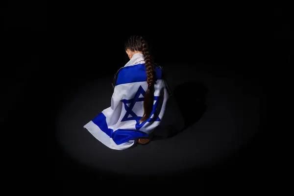 Little Jewish girl with flag of Israel sitting on black background, back view. International Holocaust Remembrance Day