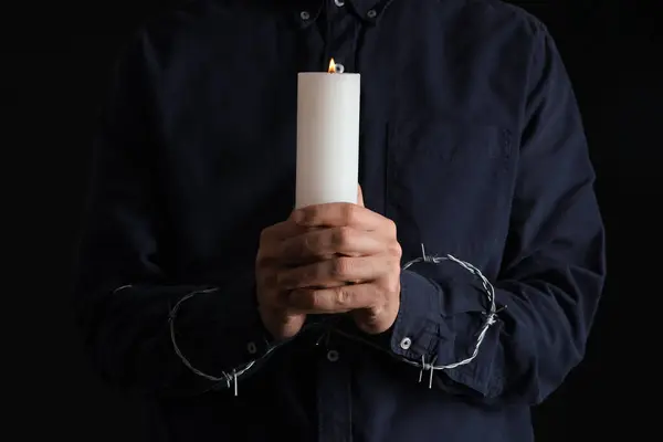 Young Jewish man with barbed wire and burning candle on black background, closeup. International Holocaust Remembrance Day