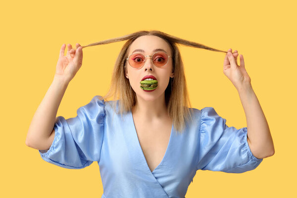 Beautiful young woman eating green sweet macaroon on yellow background