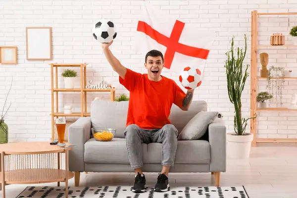 Young man with soccer balls, glass of beer, bowl of potato chips and flag of England on sofa at home