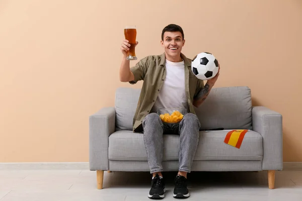 Young man with bowl of potato chips, soccer ball, glass of beer and flag of Spain on sofa near beige wall