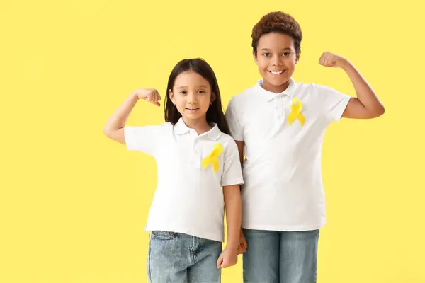 Cute little children with yellow ribbons showing muscles on color background. Childhood cancer awareness concept
